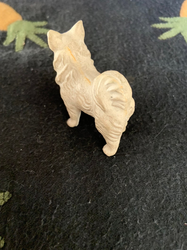Small plastic Pomeranian figurine. Vintage dog breed collectible. A white LINEOL figurine of a dog standing on a black surface. The dog is facing away from the camera and has its tail curled up.