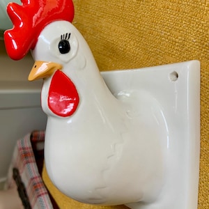 May include: A white ceramic wall-mounted chicken head sculpture featuring a bright red comb and wattle, a yellow beak, and black painted eyes with eyelashes. The chicken is attached to a square white base with a small hole for mounting purposes.