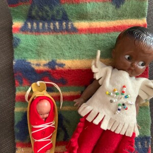 May include: Two dolls dressed in Native American clothing. The larger doll has dark skin and is wearing a white shirt with beaded trim, red pants, and fringe. The smaller doll is wrapped in a red blanket and is lying in a brown cradle.