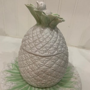 May include: A white ceramic pineapple shaped sugar bowl with a green leafy lid and a matching green and white plate. The pineapple has a detailed textured design.