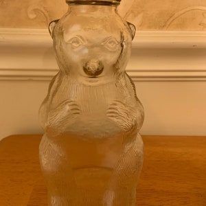 May include: A clear glass bottle shaped like a standing bear with textured fur details. The bottle features a metal screw-on cap with a red and white flame-like pattern around the rim. The bear has moulded facial features and paws.