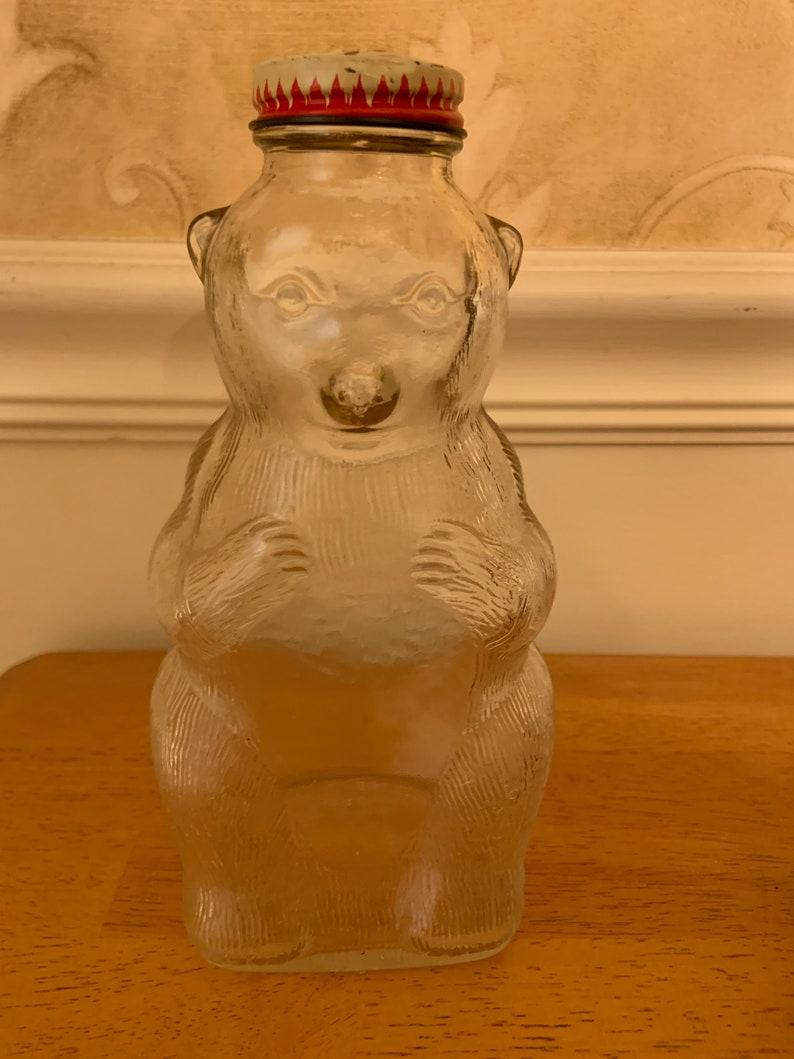 May include: A clear glass bottle shaped like a standing bear with textured fur details. The bottle features a metal screw-on cap with a red and white flame-like pattern around the rim. The bear has moulded facial features and paws.