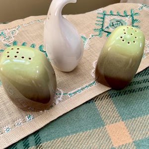 May include: A set of three ceramic kitchen items. Two salt shakers with a green and brown ombre effect and a white, curved pepper shaker. The salt shakers have multiple holes on top.