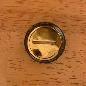 May include: A circular, metallic object with a polished gold-tone finish and a dark, weathered outer rim. The centre features a horizontal, rectangular slot cut into the surface, revealing a light-coloured interior material through the opening.