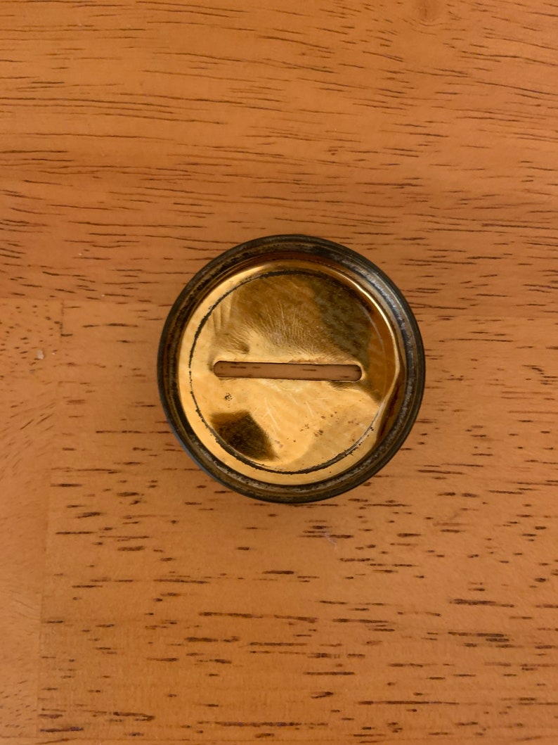 May include: A circular, metallic object with a polished gold-tone finish and a dark, weathered outer rim. The centre features a horizontal, rectangular slot cut into the surface, revealing a light-coloured interior material through the opening.