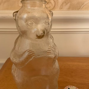 May include: A clear glass bottle shaped like a bear with textured fur details and a threaded neck opening. Beside the bottle sits a round metal lid with a coin slot, featuring the red printed text CREST BOTTLE MONS BANK around the edge.