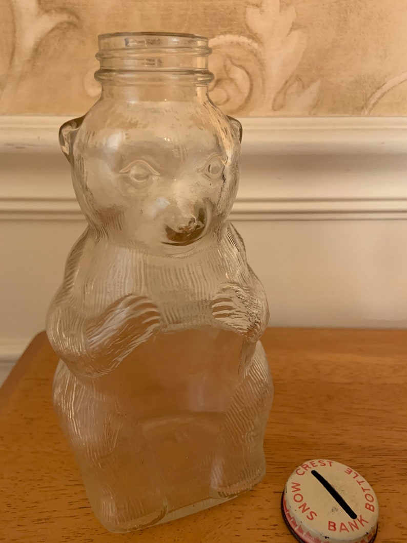 May include: A clear glass bottle shaped like a bear with textured fur details and a threaded neck opening. Beside the bottle sits a round metal lid with a coin slot, featuring the red printed text CREST BOTTLE MONS BANK around the edge.