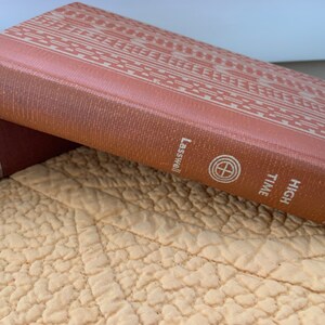 May include: Two vintage hardcover books with brown covers. The top book has a geometric pattern and the text "HIGH TIME" and "Lasswell" on the spine. The bottom book has the text "REBECCA" and "DAPHNE DU MAURIER" on the spine.