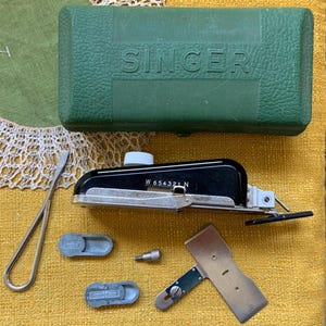May include: A collection of vintage Singer sewing machine accessories. Includes a green case with "SINGER" embossed on the lid, a black and silver attachment, small gray parts, a screwdriver, and a metal tool. The items are arranged on a yellow surface.