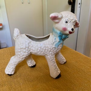 May include: A ceramic lamb planter featuring a cream-colored textured body, a light blue painted bow around its neck, pink cheeks, and black painted hooves. The hollow opening for plants is located on the back of the lamb, which stands on four legs.