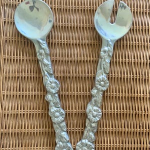 May include: A pair of silver-colored salad servers. One is a spoon, the other a fork. Both have floral and leaf detailing on the handles. The servers are set against a woven wicker background.