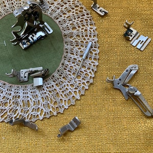 May include: A collection of silver sewing machine presser feet and a small screwdriver are arranged on a yellow textured surface. A green fabric circle is centered with a white lace doily. The presser feet are of various shapes and sizes.
