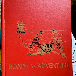May include: A bright red hardcover book titled ROADS to ADVENTURE. The front cover features a gold-colored illustration of two figures digging for a treasure chest on a beach, with a sailing ship visible in the background on the horizon.