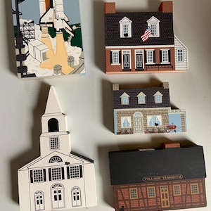 May include: A collection of painted wooden decorative items. Includes a depiction of a space shuttle launch, a house with an American flag, a church, and two other house-shaped ornaments. The items are painted in various colors.