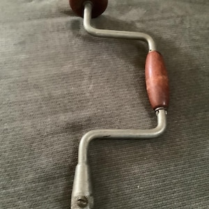May include: A vintage hand drill with a wooden handle and knob. The drill has a silver-colored metal frame and a brown wooden handle. The drill is designed for manual operation and is likely used for woodworking or other similar tasks.