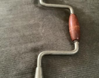 May include: A vintage hand drill with a wooden handle and knob. The drill has a silver-colored metal frame and a brown wooden handle. The drill is designed for manual operation and is likely used for woodworking or other similar tasks.