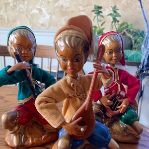 May include: Three vintage ceramic figurines of musicians. Each figure is dressed in colorful fabric clothing and playing a different instrument. The figurines have painted faces and are seated on ornate bases.