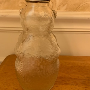 May include: A clear glass bottle shaped like a bear with a textured surface. The bottle features a metal cap with a red and white flame-like pattern around the rim. The glass body has a rounded silhouette with small ears at the top of the neck.