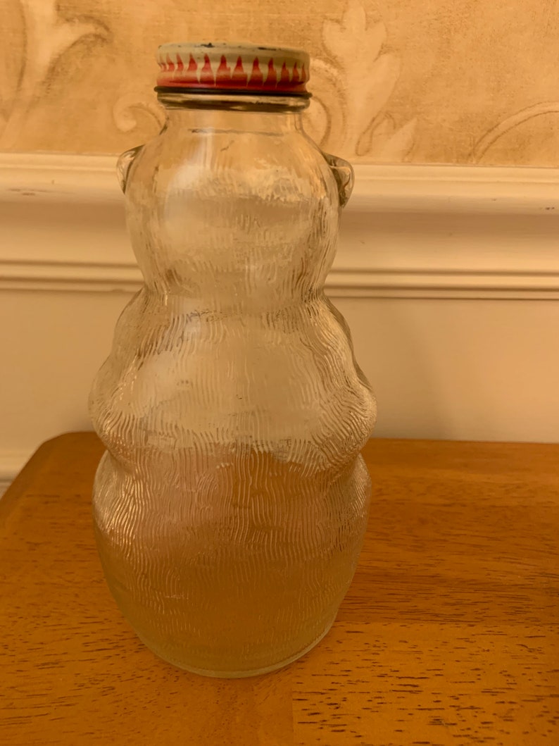 May include: A clear glass bottle shaped like a bear with a textured surface. The bottle features a metal cap with a red and white flame-like pattern around the rim. The glass body has a rounded silhouette with small ears at the top of the neck.