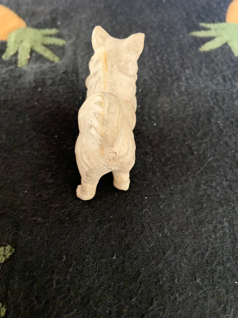 A white LINEOL figurine of a dog standing on a black surface. The dog is facing away from the camera and has a long tail.