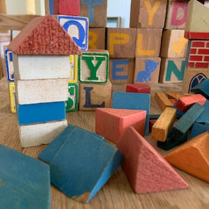 May include: A collection of colorful wooden building blocks, including cubes with letters and numbers, and triangular and rectangular shapes. The blocks are in various colors, including red, blue, yellow, and green. Some blocks are stacked to resemble houses.