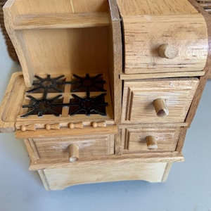 May include: A miniature wooden stove with a light brown finish. The stove features a four-burner cooktop with black grates, multiple drawers, and a small oven door. The knobs and handles are also made of wood.