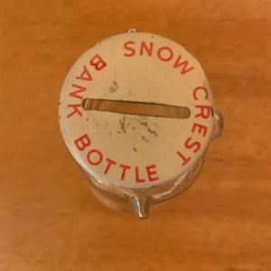 May include: A clear glass bottle with a round, off-white metal lid featuring a horizontal coin slot. The lid is printed with the red text SNOW CREST BANK BOTTLE arranged in a circular pattern around the centre slot, showing minor surface wear.