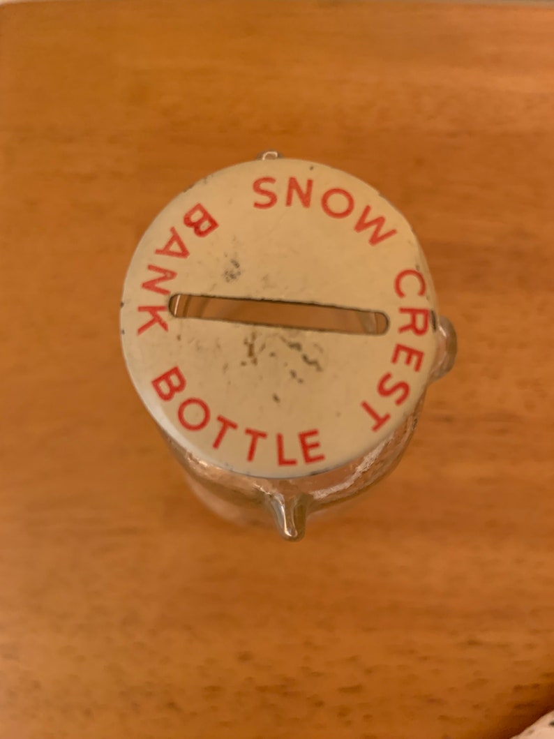 May include: A clear glass bottle with a round, off-white metal lid featuring a horizontal coin slot. The lid is printed with the red text SNOW CREST BANK BOTTLE arranged in a circular pattern around the centre slot, showing minor surface wear.