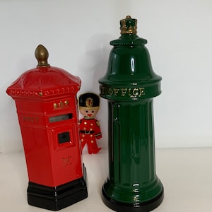 May include: Two ceramic piggy banks shaped like a red British post box and a green post office pillar box. The post box has a slot for mail and the words "POST" and "G.R." on the front. The post office pillar box has a gold crown on top and the words "POST OFFICE" on the front.