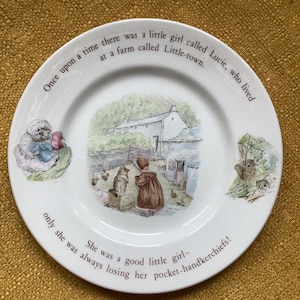 May include: White ceramic plate with a detailed illustration of a girl, cat, and farm scene. The plate has text around the rim that reads, "Once upon a time there was a little girl called Lucie..." and "...always losing her pocket-handkerchiefs!"