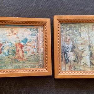 May include: Two framed vintage religious art prints. The prints feature detailed scenes with figures in classical attire, set against a backdrop of landscapes and sky. The frames are made of light brown wood with decorative edges.