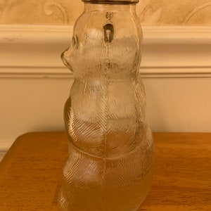 May include: A clear glass bottle shaped like a bear with textured fur details. The bottle features a metal screw-top lid with a red and white decorative pattern around the rim. The glass is transparent and moulded into the form of a seated bear.