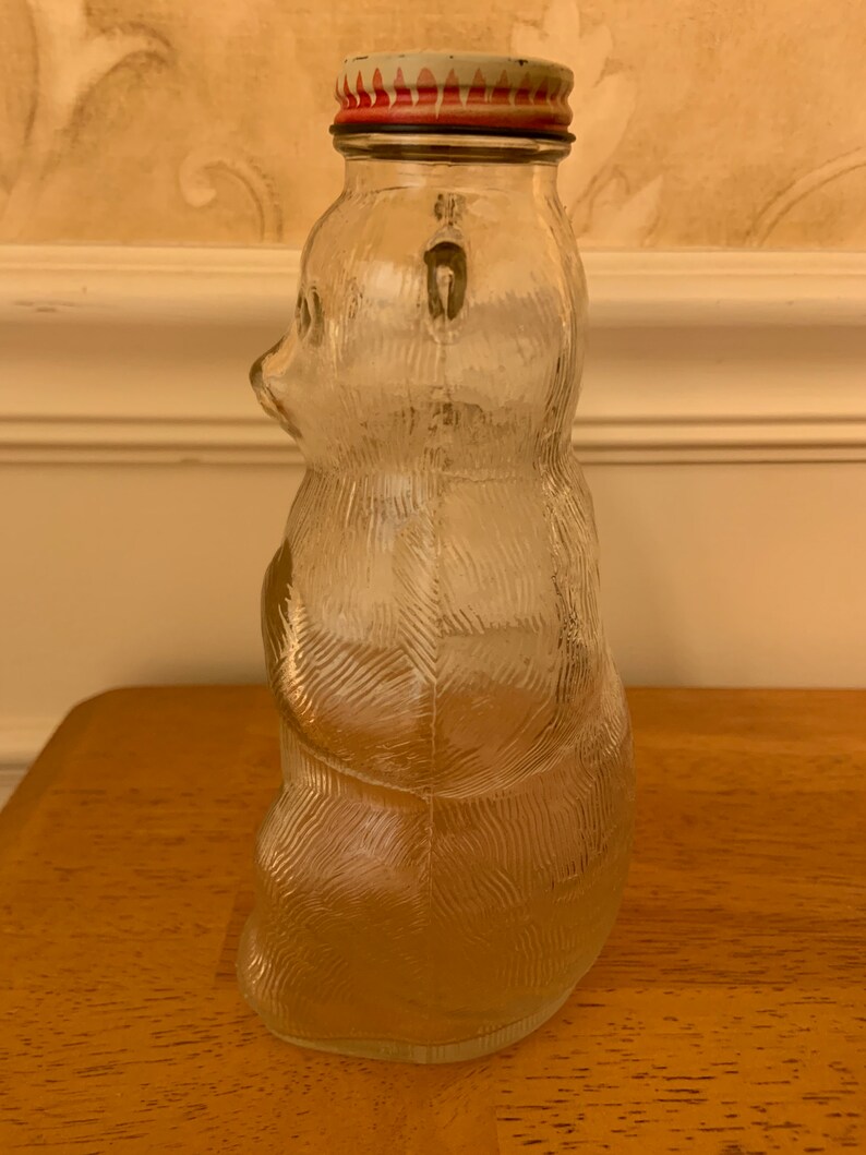 May include: A clear glass bottle shaped like a bear with textured fur details. The bottle features a metal screw-top lid with a red and white decorative pattern around the rim. The glass is transparent and moulded into the form of a seated bear.