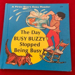 May include: A vintage children's book cover with a yellow background and blue border. The title is "The Day Busy Buzzy Stopped Being Busy". The cover features a boy holding a fishbowl with a goldfish inside, and a dog looking at the boy.