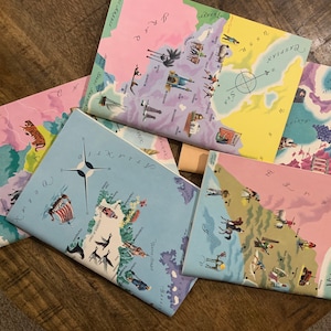 May include: Five colorful illustrated maps with a compass and various images of animals, people, and places. The maps are folded and arranged on a wooden surface.