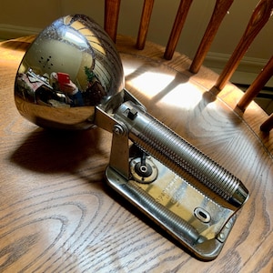 May include: A vintage chrome-plated spotlight with a ribbed handle and a pivoting base. The spotlight has a large, reflective bulb housing and is labeled "Sportsman" on the base. It is resting on a wooden chair.