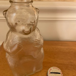 May include: A clear glass bear-shaped bottle with textured fur details and a narrow neck. Beside the bottle sits a round metal lid featuring a red serrated edge and the text SNOW CREST BANK BOTTLE printed in red around a central coin slot.