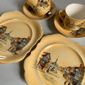 May include: A vintage tea set with a cream-colored background. The set includes two teacups with saucers and two plates. Each piece features a detailed illustration of a town scene with buildings and people. The edges of the plates have a scalloped design.