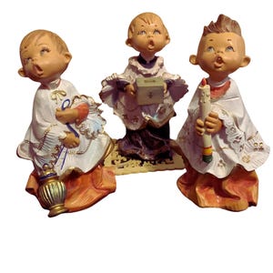 May include: Three ceramic figurines of choirboys in white robes with gold and orange accents. One holds a lantern, another a box, and the third a candle. The figurines have detailed facial features and are posed with their heads tilted upwards.
