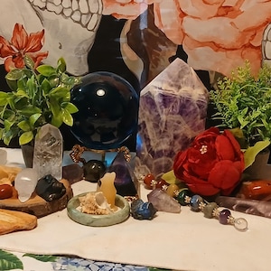 May include: A collection of crystals and stones including a dark blue sphere, a large amethyst point, a clear quartz point, a green stone ring, a red flower, and two small potted green plants arranged on a white cloth with a floral background.