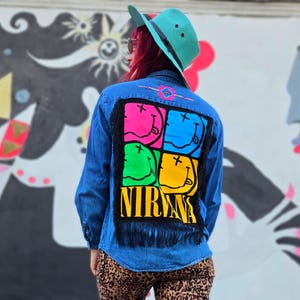 May include: Blue denim jacket with a colorful Nirvana smiley face design on the back. The design features four squares with smiley faces in pink, blue, green, and yellow, with the band's name below. The jacket has black fringe.