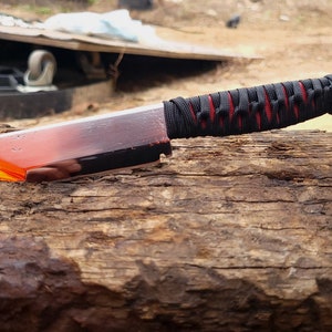 May include: A fixed-blade knife with a metallic silver blade featuring a bright orange translucent section near the tip. The handle is wrapped in black cord over a red base, and the knife is balanced horizontally on a metal nail.