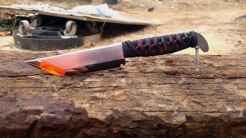 May include: A fixed-blade knife with a metallic silver blade featuring a bright orange translucent section near the tip. The handle is wrapped in black cord over a red base, and the knife is balanced horizontally on a metal nail.