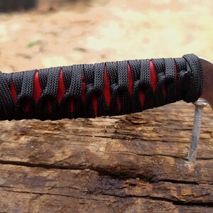 May include: A knife handle wrapped in black cord over a red base, featuring a curved metal pommel at the end. The handle is positioned horizontally, showcasing the intricate crisscross pattern of the black cord wrapping against the red material.