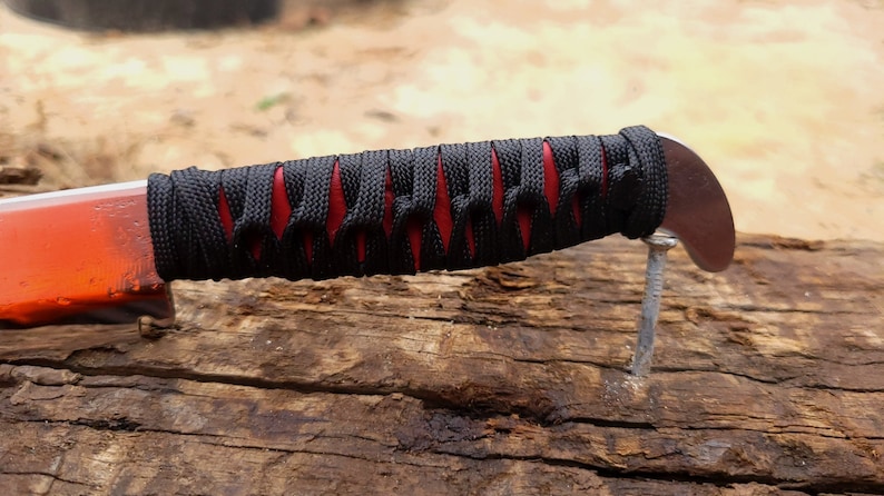 May include: A knife handle wrapped in black cord over a red base, featuring a curved metal pommel at the end. The handle is positioned horizontally, showcasing the intricate crisscross pattern of the black cord wrapping against the red material.