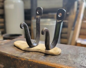 May include: A double wall hook forged from dark gray metal, featuring two upward-curving prongs with curled tips and circular mounting holes at the top of each vertical arm, set upon a light-colored, textured, elongated oval base plate.