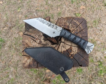 May include: A fixed-blade knife with a silver-toned, textured steel blade featuring etched runic symbols and a black cord-wrapped handle. The knife includes a black leather sheath embossed with a hammer design and a metal snap closure.