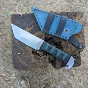 May include: A fixed-blade knife with a silver tanto-style steel blade, a black handle wrapped with teal accents, and a rounded metal pommel. Beside the knife is a blue leather sheath featuring two black leather straps and a metal snap button.