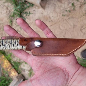 May include: A fixed-blade knife featuring a handle wrapped in a cream-colored cord with black accents and a metallic pommel. The blade is enclosed in a brown leather sheath secured with a silver-colored metal snap button fastener.
