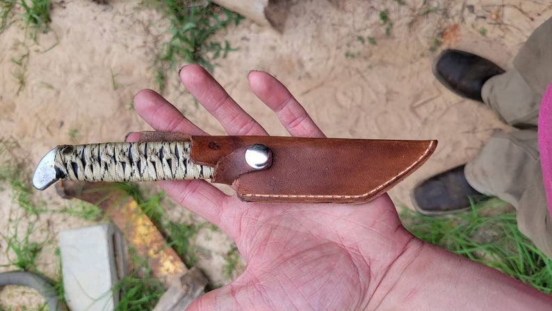 May include: A fixed-blade knife featuring a handle wrapped in a cream-colored cord with black accents and a metallic pommel. The blade is enclosed in a brown leather sheath secured with a silver-colored metal snap button fastener.